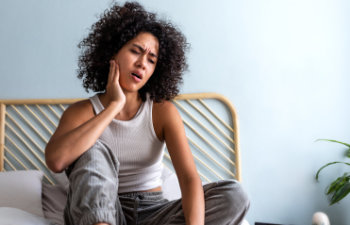 young woman with curly hair touching cheek in pain having toothache sitting on bed, Lawrenceville, GA