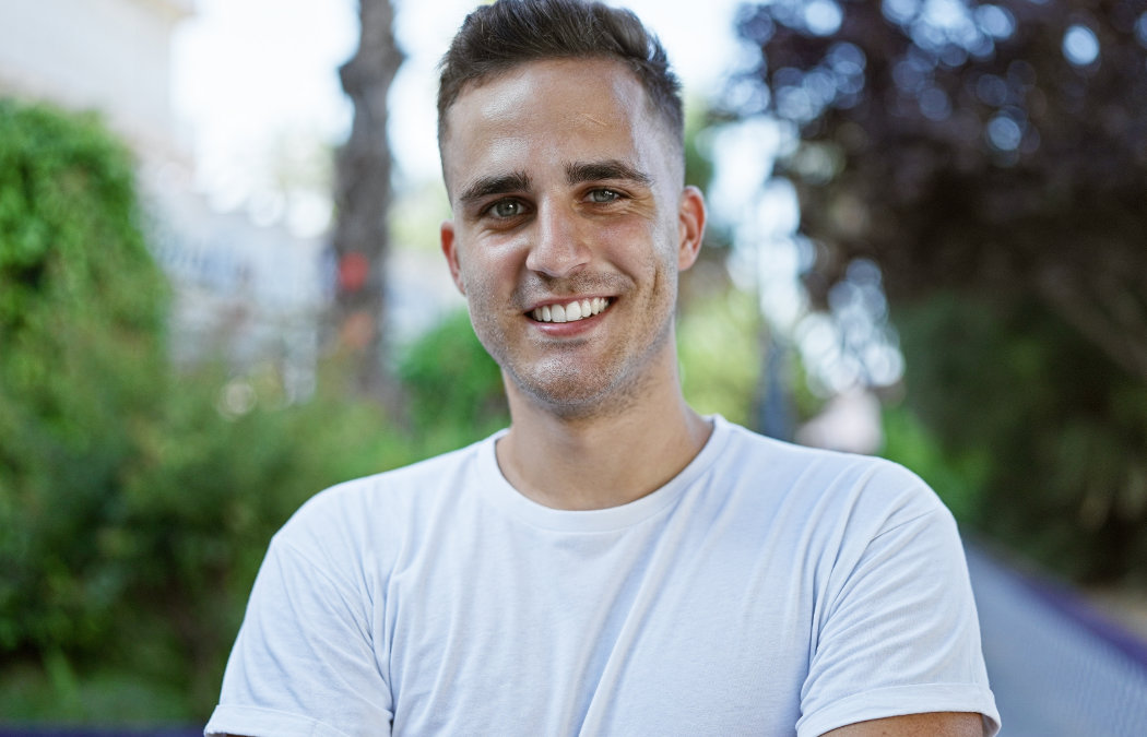 A man wearing a white t-shirt stands outdoors, smiling at the camera, with greenery and trees in the blurred background., Lawrenceville, GA