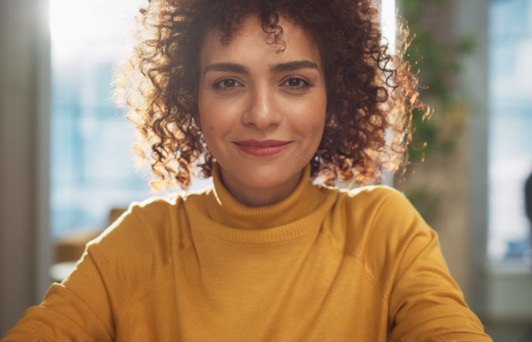 A person with curly hair and a yellow turtleneck sweater smiles while sitting indoors with sunlight in the background., Lawrenceville, GA