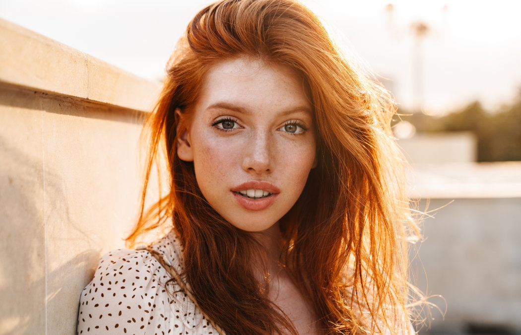 A young woman with long red hair and freckles looks directly at the camera, standing outdoors in warm sunlight next to a concrete wall., Lawrenceville, GA