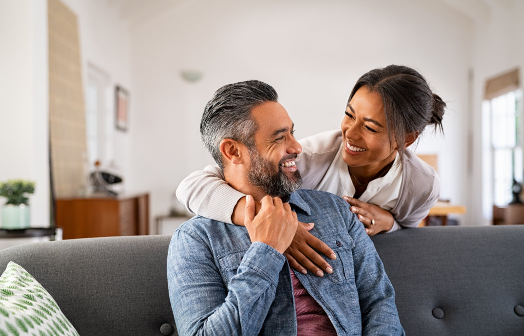 A man sits on a couch smiling at a woman who stands behind him with her arm on his shoulder; both appear happy in a bright living room., Lawrenceville, GA